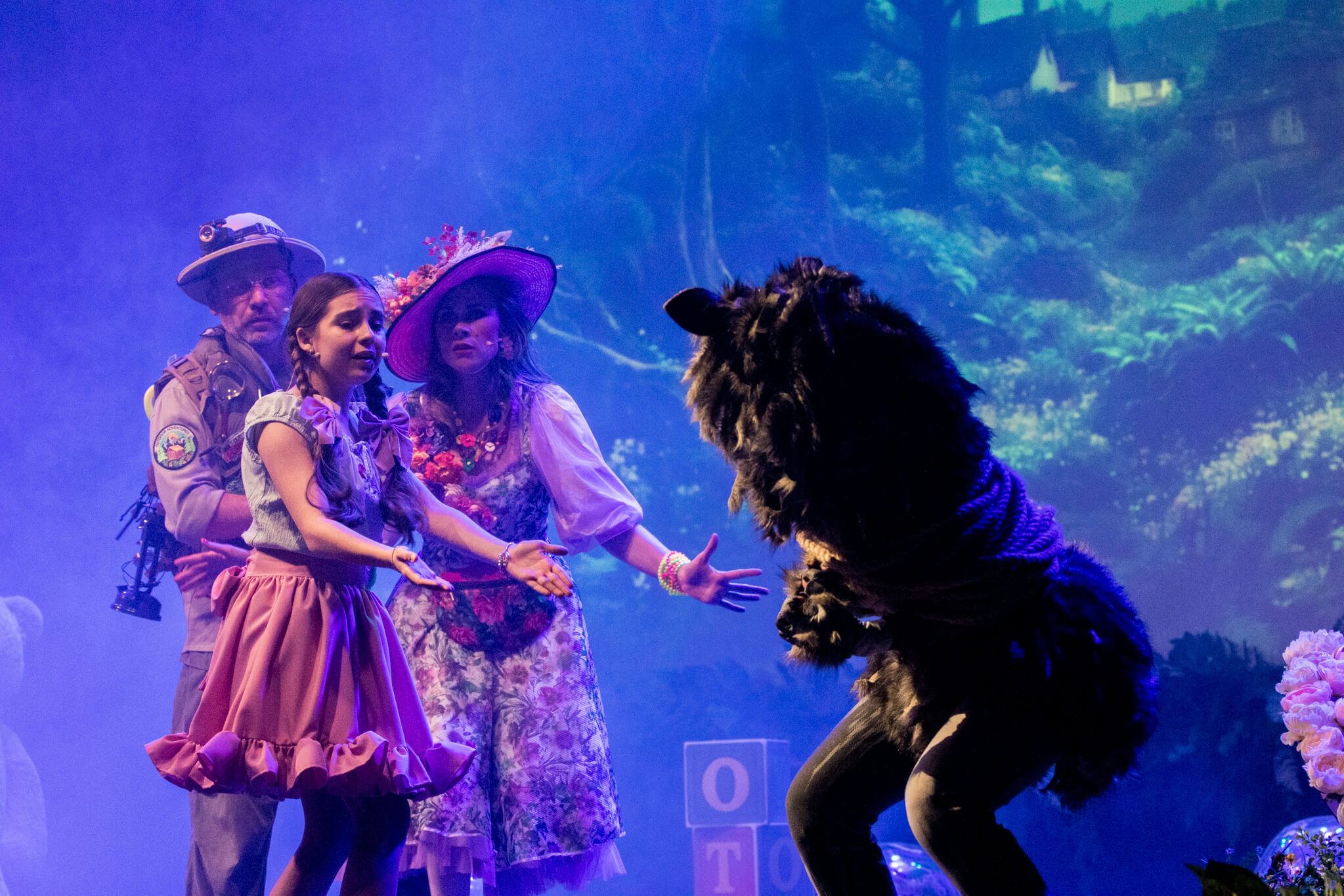 Grupa postaci na scenie – dziewczynka w różowej sukience, mężczyzna w mundurze i kobieta w kwiatowej sukience, stoją w pobliżu wilka w futrzanym kostiumie. W tle fantastyczna sceneria leśna.