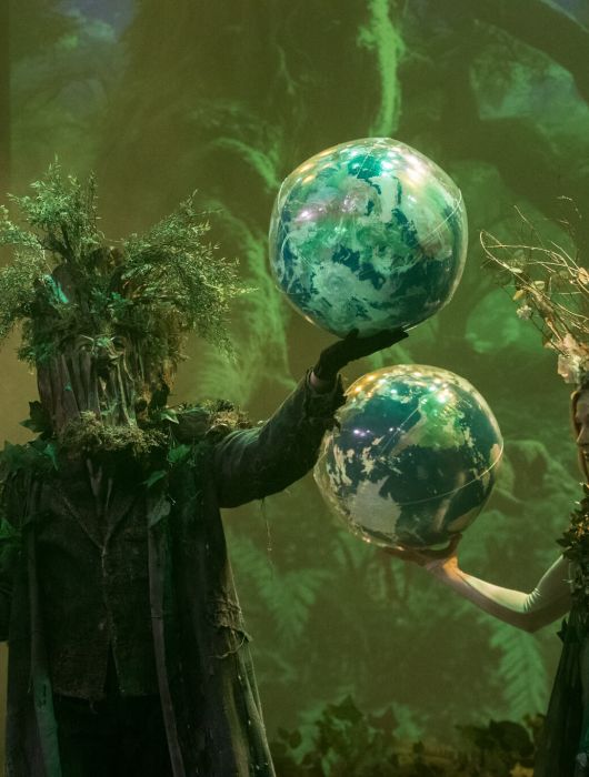 Dwie postacie w zielonej scenografii trzymają podświetlane kule symbolizujące planety.
