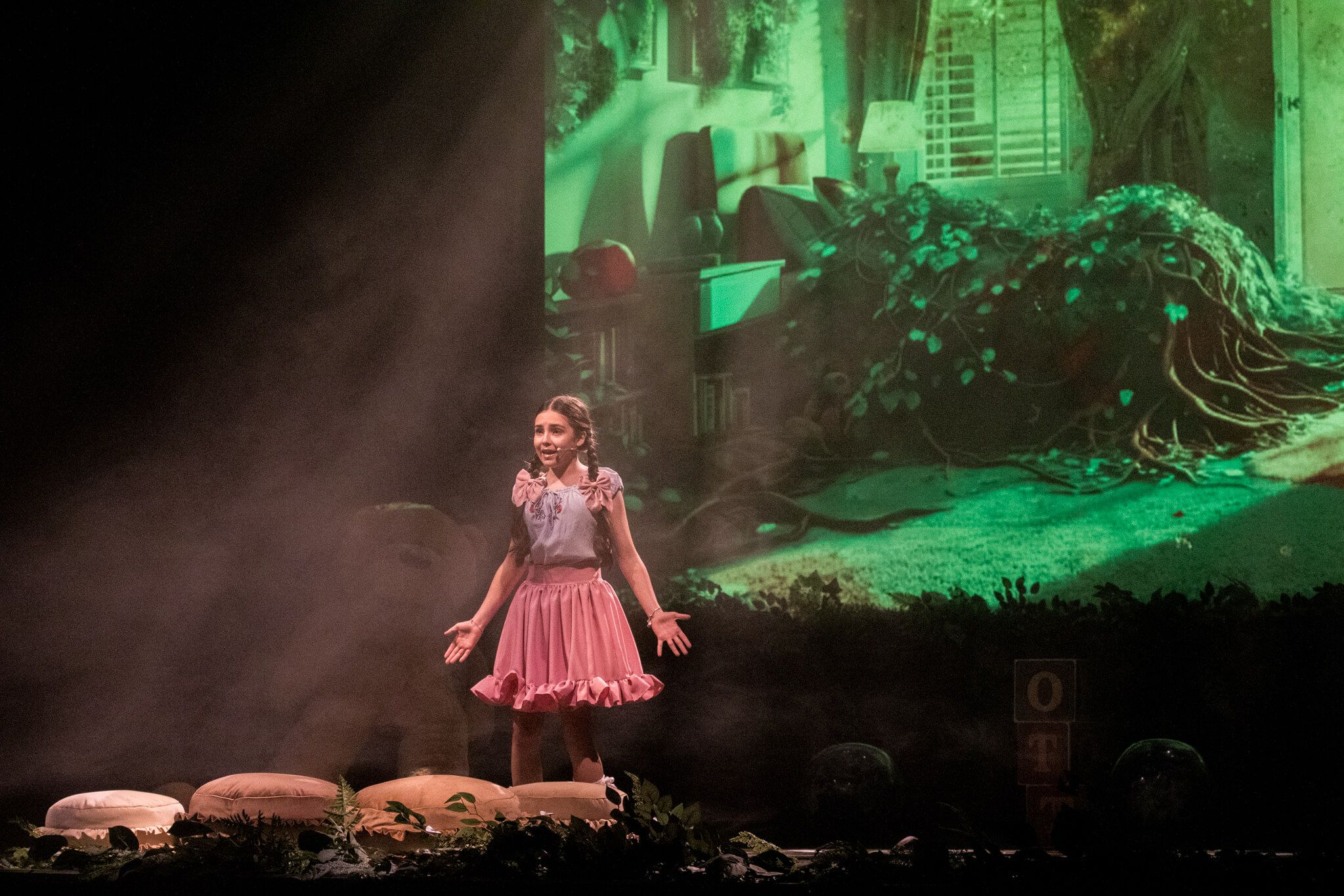Dziewczynka w różowej sukience stoi na scenie, gestykulując, jakby mówiła lub śpiewała. Tło to zaciszne wnętrze z roślinami i dużym misiem. Na scenie widać dym i tajemniczy nastrój.