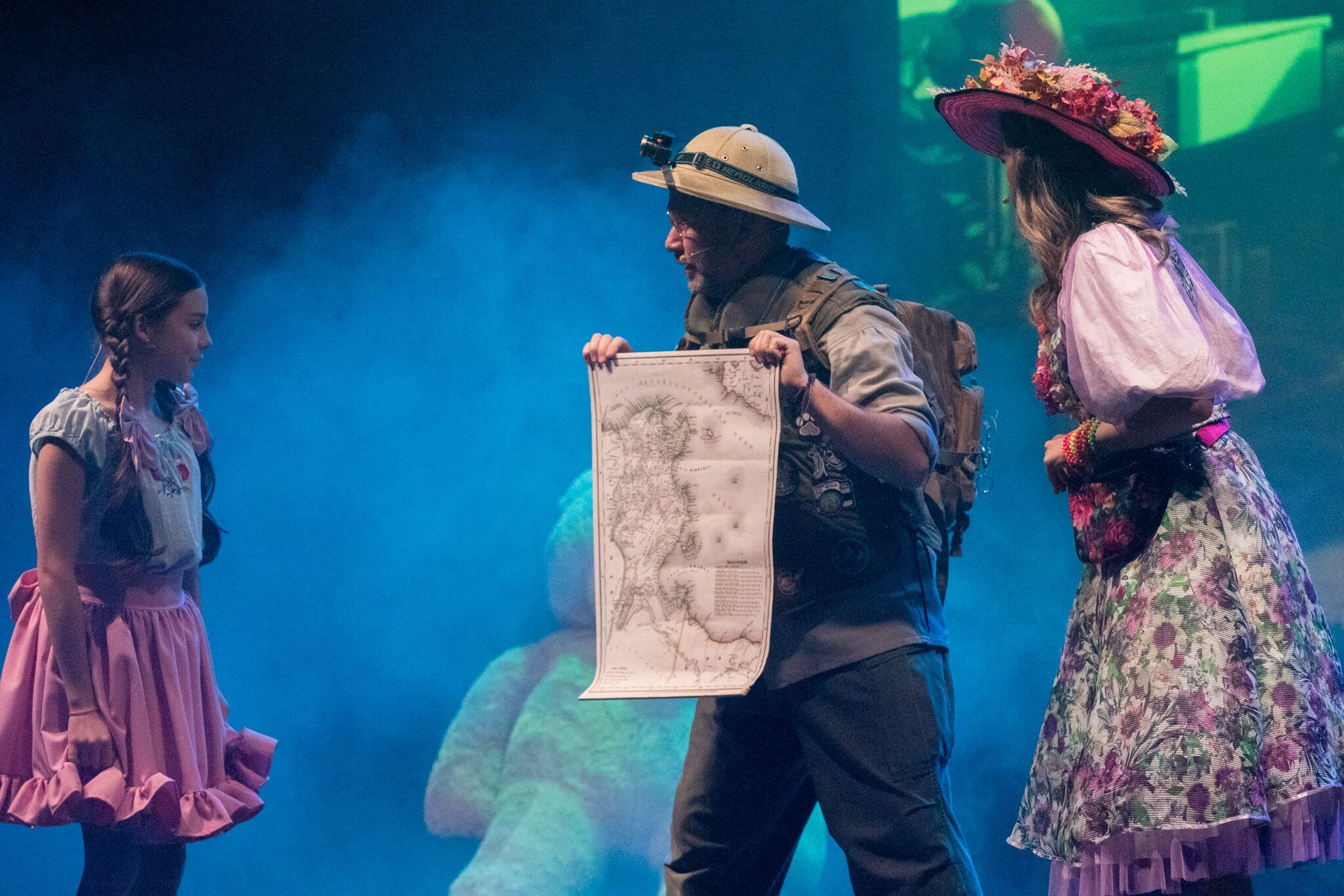 Na scenie mężczyzna, który w stroju podróżnika pokazuje mapę dla dziewczynki i kobiety.