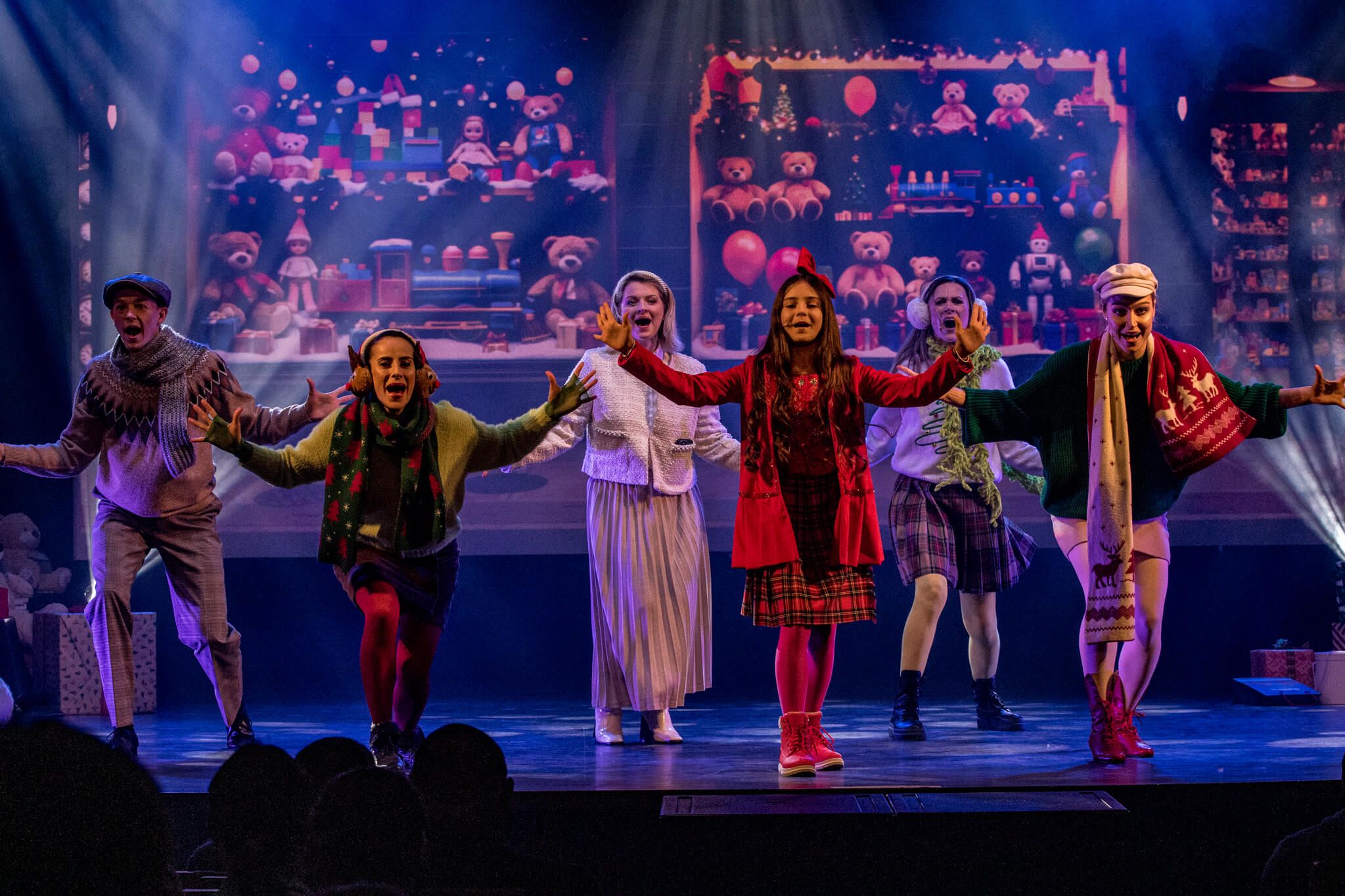 Sześcioro aktorów w kolorowych zimowych kostiumach z rozłożonymi ramionami na scenie