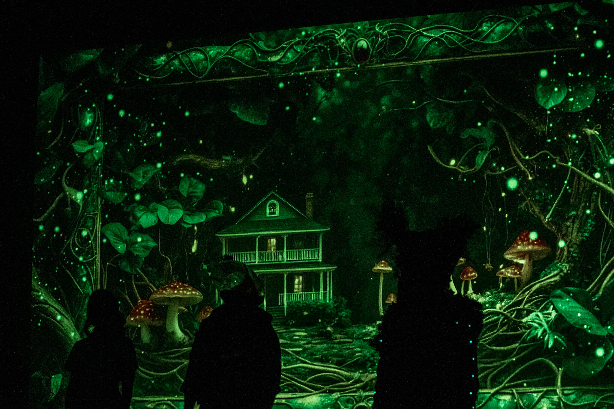 Trzy postacie na tle scenografii z dużym domem, grzybami i świetlistymi punktami, symbolizującej tajemniczy las.