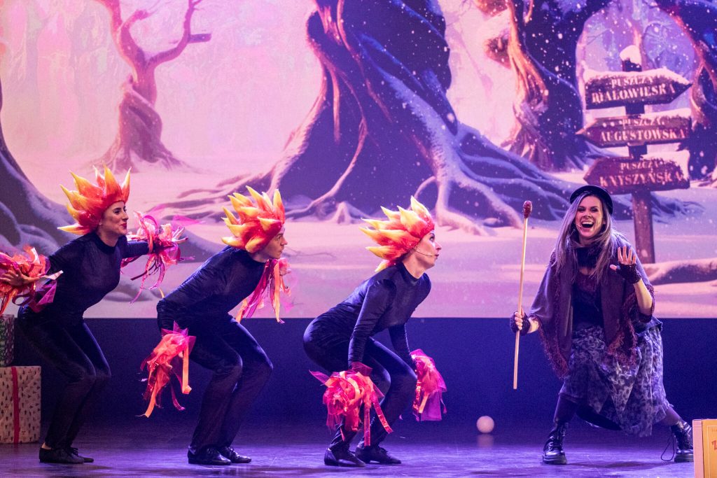 Trzech tancerzy w czarnych strojach z ognistymi ozdobami i postać czarownicy na tle zimowej scenerii
