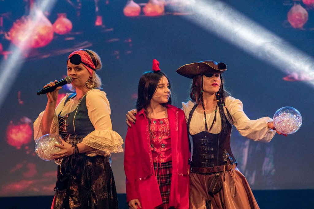 Dwie piratki i dziewczynka w czerwonej sukience na scenie z podwodnym tłem z meduzami
