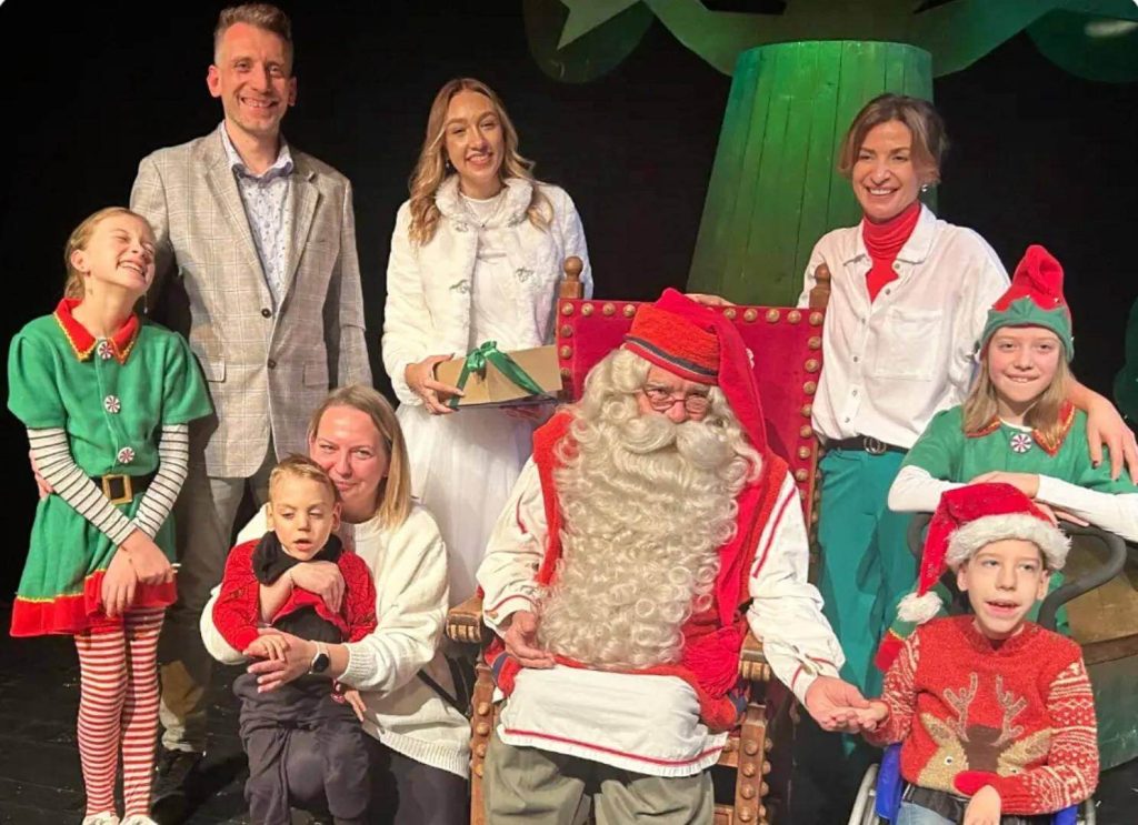 Mikołaj i aktorzy w świątecznych kostiumach stoją na oświetlonej scenie.