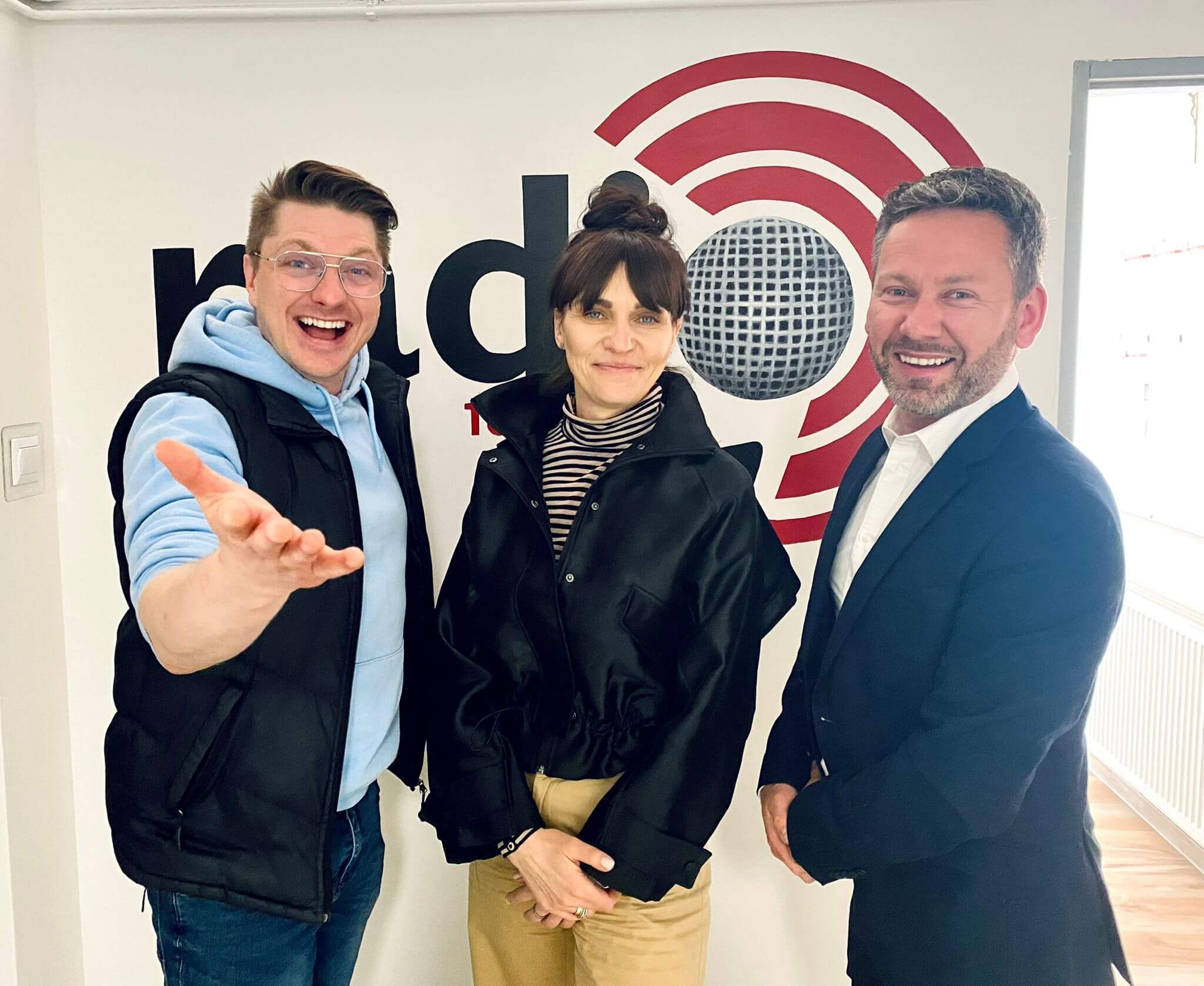 Trzy osoby stojące przed ścianą z logo stacji radiowej